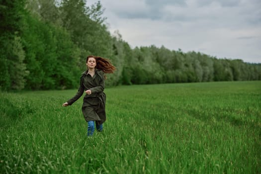 a beautiful woman in a long raincoat runs across a field in high grass in spring in cloudy weather