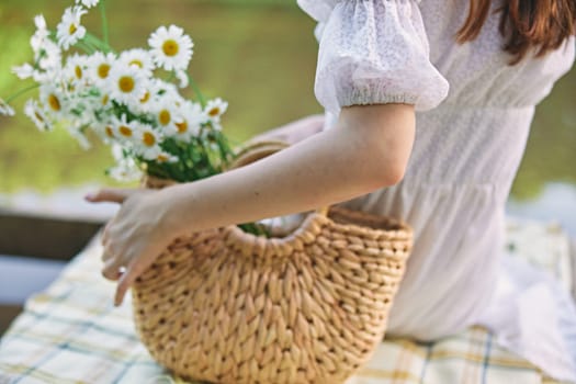 a woman sits with her back to the camera by the lake holding a wicker basket with daisies in her hand. subject photography without a face