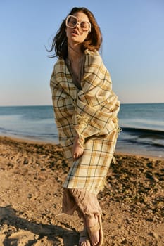 a woman wrapped in a plaid stands on the seashore in bright sunglasses and squints from the sun