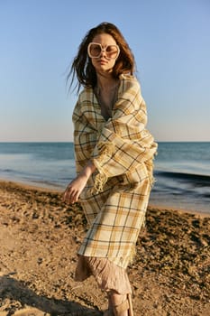a woman wrapped in a plaid stands on the seashore in bright sunglasses and squints from the sun