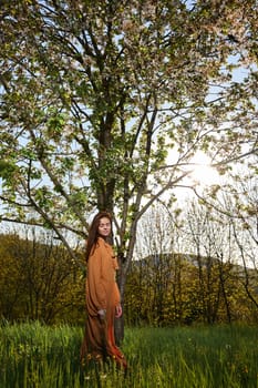 a slender, calm woman with long red hair stands in the countryside near a flowering tree in a long orange dress and looks into the camera enjoying the silence