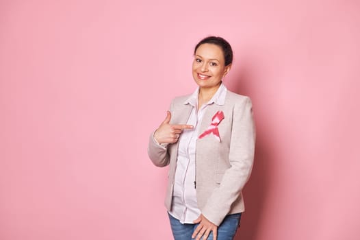 Positive multi-ethnic woman, standing over pink background and pointing her finger at a pink satin ribbon on her jacket