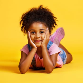 Smile, cute and portrait of a child on the floor isolated on a yellow background in a studio. Adorable, sweet and an innocent girl lying down looking happy, beautiful and relaxed on a backdrop