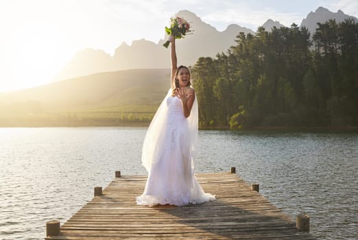 Wedding, bouquet and celebration with a bride on a pier over a lake in nature after a marriage ceremony. Happy, smile and flowers with a woman celebrating being married in the tradition of matrimony