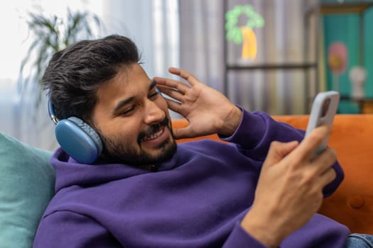 Happy indian man in wireless headphones relaxing sitting on sofa at home listening favorite music