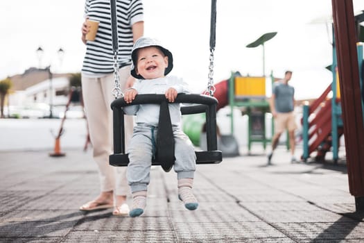Mother pushing her infant baby boy child on a swing on playground outdoors.