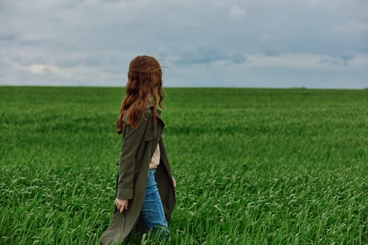 a red-haired woman in a long raincoat stands in a green field in cloudy weather in a strong wind