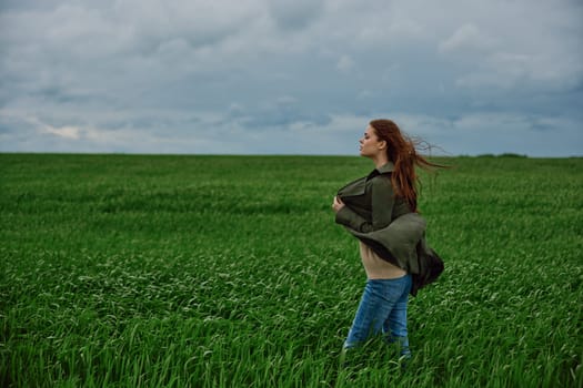 a red-haired woman stands in a green field in rainy, cold weather, holding a raincoat in the wind