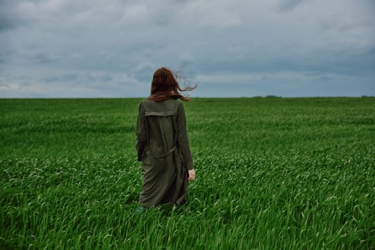 a woman in a long raincoat stands in a green field in windy weather with her back to the camera. High quality photo