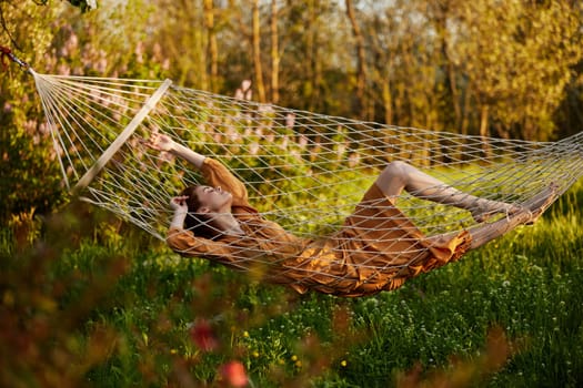 a beautiful woman is resting in nature lying in a mesh hammock in a long orange dress looking to the side, arms outstretched. Horizontal photo on the theme of recreation