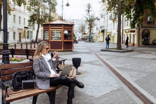 Business adult caucasian woman sitting with laptop and talking on speakerphone in city square