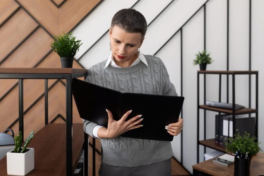 a businesswoman with a short haircut in a gray sweater stands in the office with a leather folder for documents and looks into it