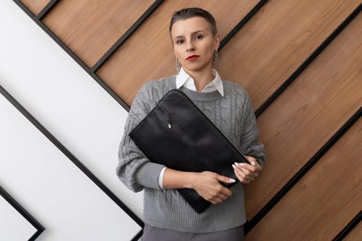 A business woman with a short haircut stands with a folder for documents on the background of a wall in the office