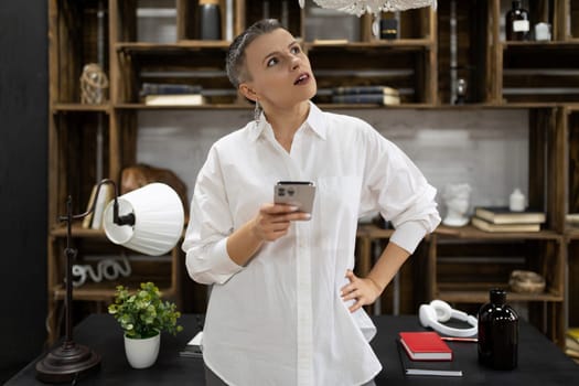 stylish mature businesswoman at the desk with a phone in her hands looks up in thought
