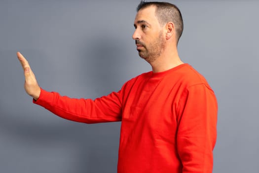 Stop. Side view of a serious bossy dark-haired man in a red pullover, prohibition gesture, saying no, empty copy space on the left for text. Studio shot isolated on gray background.