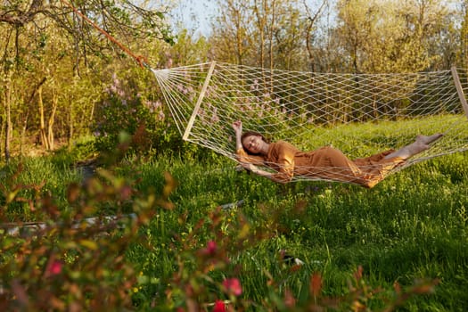 a happy woman in a long orange dress is relaxing in nature lying in a mesh hammock enjoying the peace and tranquility in the country surrounded by green foliage