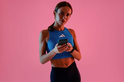 Sporty woman holding smartphone standing against studio background and looking at camera