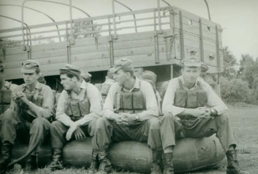 Retro photo shows young men - soldiers during summer time. Soldiers pose with swimming belts - vest and sit on the rubber boat. Vintage photography. Circa 1970.