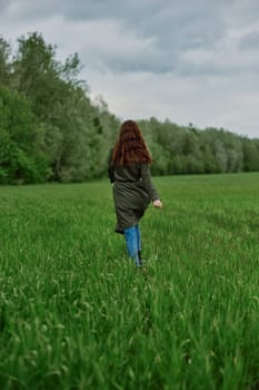 a woman in a long raincoat runs across a field in tall green grass in cloudy weather in spring