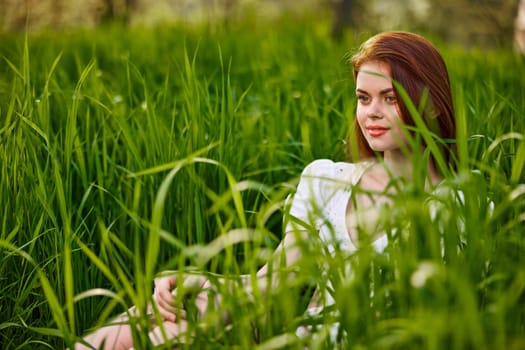 a pretty woman straightens her red hair while sitting in the tall grass in a light dress