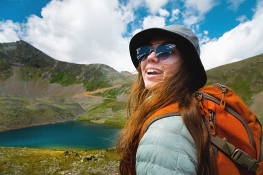 portrait of a girl in the mountains standing looking at the mountain landscape. Happy woman in a cap enjoys the sunshine while hiking in the mountains. Tourist with glasses