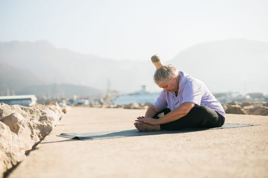 Mature woman with dreadlocks working out doing yoga exercises on sea beach copy space - wellness well-being and active elderly age concept