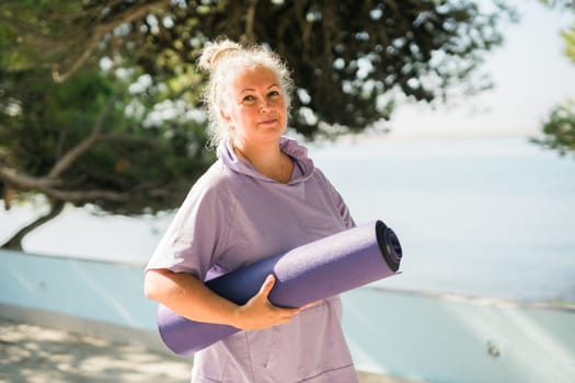 Old woman holding yoga mat and preparing to practice yoga or meditation outdoors on sea beach. Happy mature woman with dreadlocks exercising on seashore. Meditation or yoga and relaxation concept