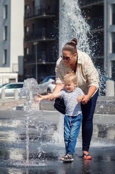 Mom and her son, in the city center, by a fountain of water. The child is afraid to touch the water so that it does not get wet, the mother encourages it
