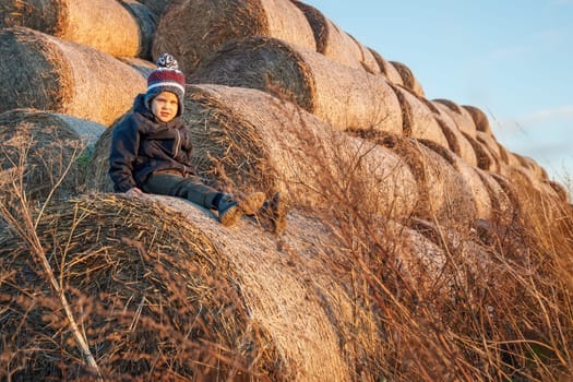 A cute little boy in autumn clothes and a knitted hat poses against a background of golden hay bales. Horizontal photo