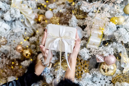 Female hands hold a gift box tied with a ribbon on the background of a Christmas tree, the concept of christmas and gifts.
