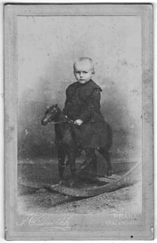 Vintage cabinet card shows portrait of the small child. Little boy sitting on rocking horse. Edwardian fashion. Photo was taken in a photo studio. Photo was taken in Austro-Hungarian Empire or also Austro-Hungarian Monarchy