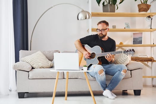 Using laptop. Man in casual clothes and with acoustic guitar is indoors