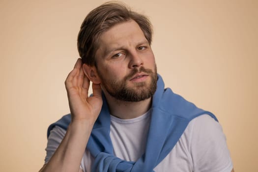 Confused man trying to hear you, frowning, keeping arm near ear for louder voice, ask to repeat