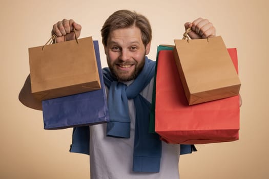 Shopaholic man showing shopping bags, advertising discounts, smiling looking amazed with low prices, shopping on Black Friday holidays. Young guy celebrate win isolated on beige studio background