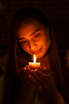 A woman holds a burning candle in her palms on a dark black background