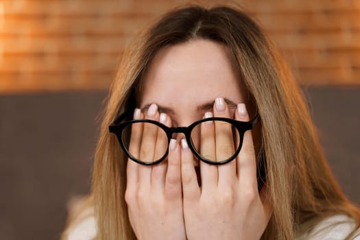 A young woman in glasses rubs her eyes with her hands.