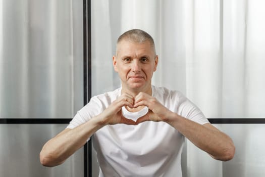 A middle-aged man in a white T-shirt smiles and shows a heart symbol