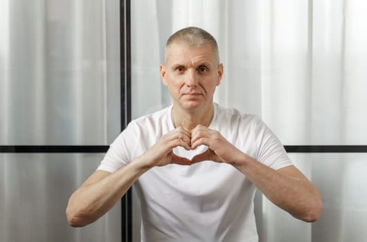 A middle-aged man in a white T-shirt smiles and shows a heart symbol