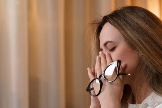 Young woman rubs her eyes, suffers from eye fatigue, holding glasses in her eyes.