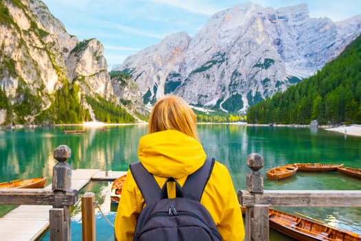 Backview of the woman with a backpack and wearing a yellow sport jacket having fun on lake Braies with turquoise water and wooden boats in the Dolomites.