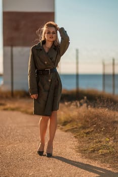 Portrait blonde sea cape. A calm young blonde in a khaki raincoat stands on the seashore against the backdrop of a lighthouse.