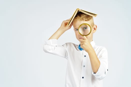 The schoolboy put the book on his head and looks through a magnifying glass. Isolated background.