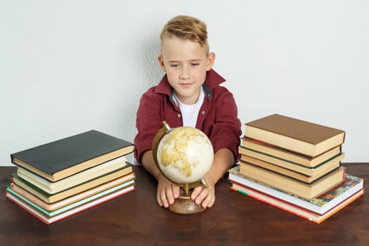 The student sits at the table and looks at the globe holding on to it with his hands. On the table there are books, a globe and an alarm clock.
