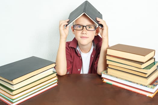 A pupil boy sits at a table with books, holding an open book on his head.