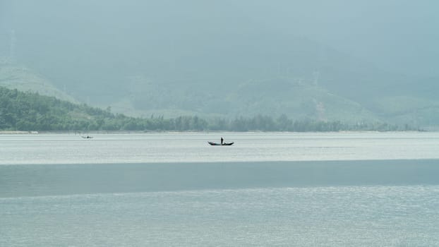 A boat with a fisherman in the middle of the river against the backdrop of foggy mountains