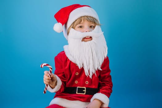 Funny boy in Santa Claus hat and sweet caramel cane on blue studio background