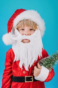 Friendly little Christmas boy in Santa Claus costume and tree on blue background