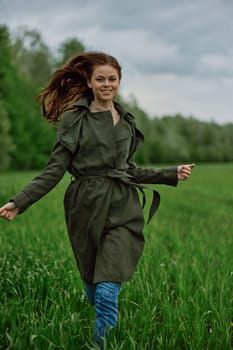 a beautiful woman in a long raincoat runs across a field in high grass in spring in cloudy weather