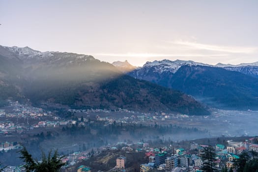 monsoon clouds moving over snow covered himalaya mountains with the blue orange sunset sunrise light with town of kullu manali valley at the base of mountains