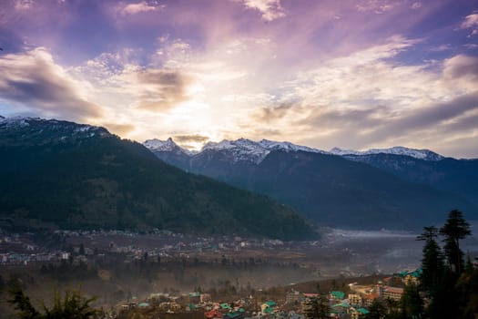 monsoon clouds moving over snow covered himalaya mountains with the blue orange sunset sunrise light with town of kullu manali valley at the base of mountains
